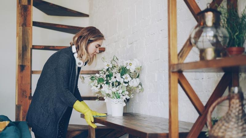 A cleaner in yellow gloves wiping a wooden table beside a vase of flowers